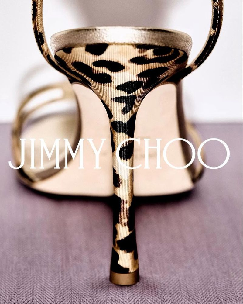 Close-up studio shot of a Jimmy Choo high heel sandal with a leopard print pattern, gold straps, and a tan footbed on a grey surface.