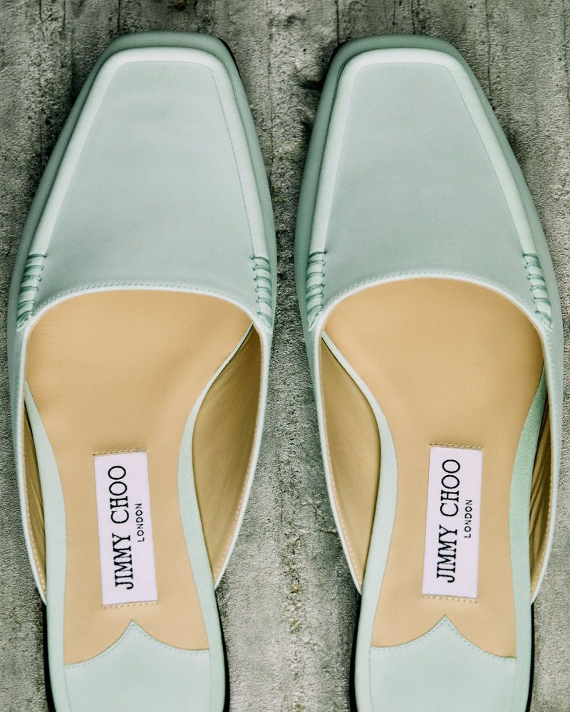 Close-up of two Jimmy Choo light blue, leather heeled mules with a square toe on a textured grey surface. Fashion footwear.