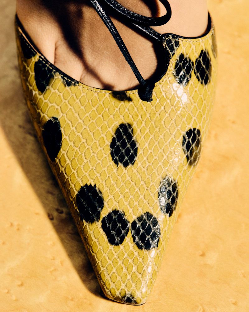 Close-up of a woman's foot wearing a yellow snakeskin-print high heel with a lace-up closure against a marble background.