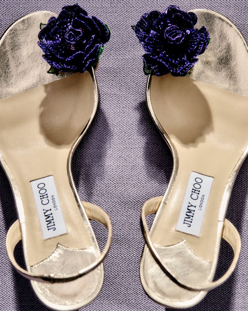 Gold Jimmy Choo sandals with purple floral embellishments, displayed on a textured grey background. Fashionable footwear close-up.