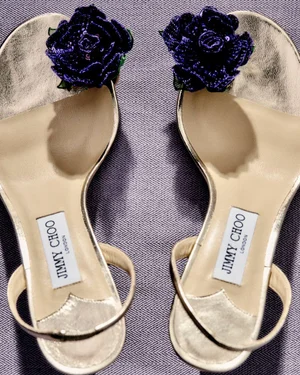 Pair of Jimmy Choo metallic slingback sandals featuring dark purple sequin floral rosettes on a textured woven surface.