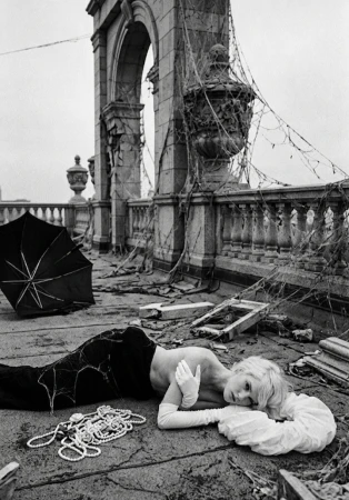 Black and white photo of a blonde woman lying on a debris-strewn stone rooftop wearing opera gloves beside pearl necklaces.