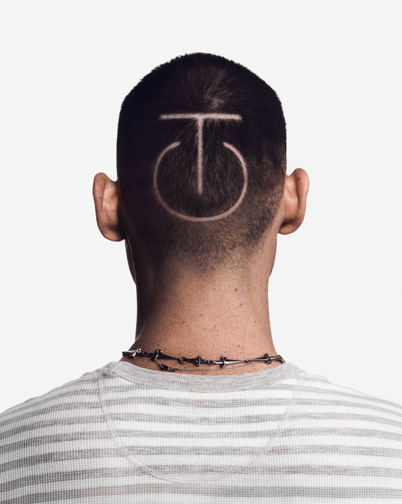 Back of a person's head with a buzzcut featuring a shaved geometric symbol, wearing a striped shirt against a white backdrop.