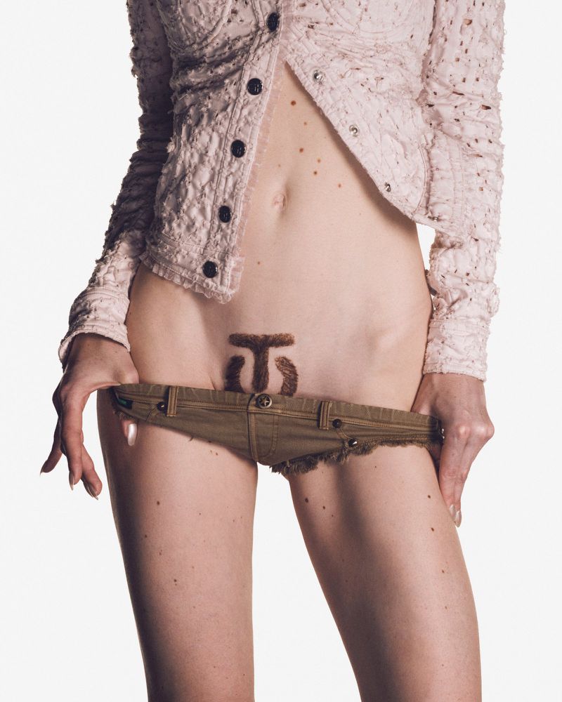 Model Lidia Judickaite in an unbuttoned textured cardigan pulling down ultra low-rise khaki shorts against a white backdrop.