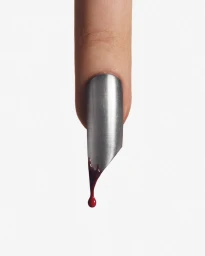 A finger with a long metallic silver pointed nail dripping a single droplet of crimson liquid against a plain white background.