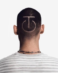 Back of a person's head with a buzzcut featuring a shaved geometric symbol, wearing a striped shirt against a white backdrop.