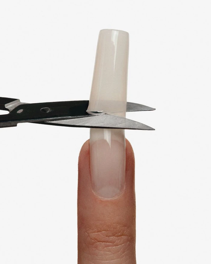 Scissors cutting an extra-long translucent acrylic nail extension on a single fingertip against a white studio background.