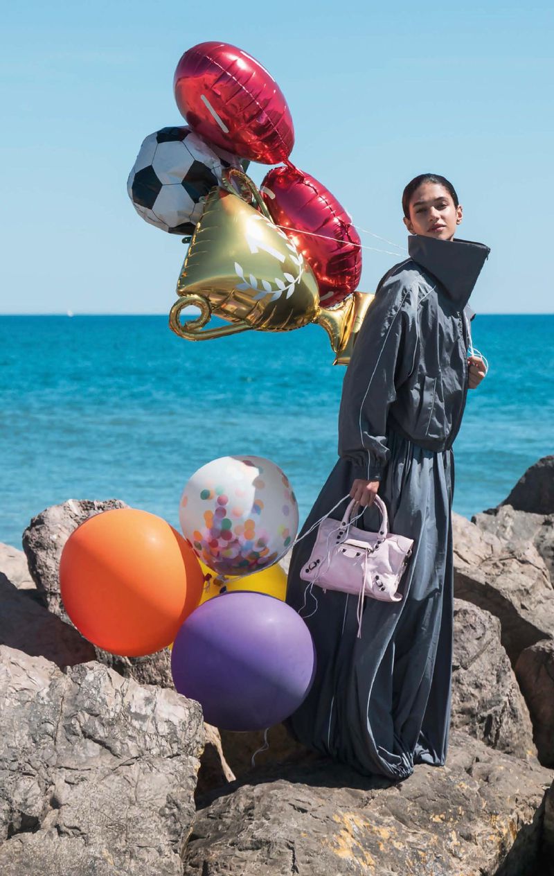 Model Tamar David standing on coastal rocks holding a cluster of colorful balloons and a pink handbag by the ocean.
