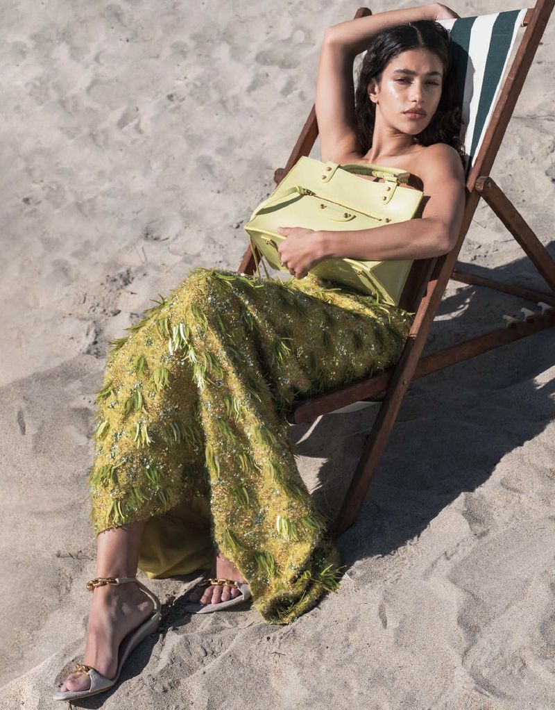 Model Tamar David reclining in a wooden deckchair on a sandy beach holding a pale yellow handbag and wearing a chartreuse fringed dress.
