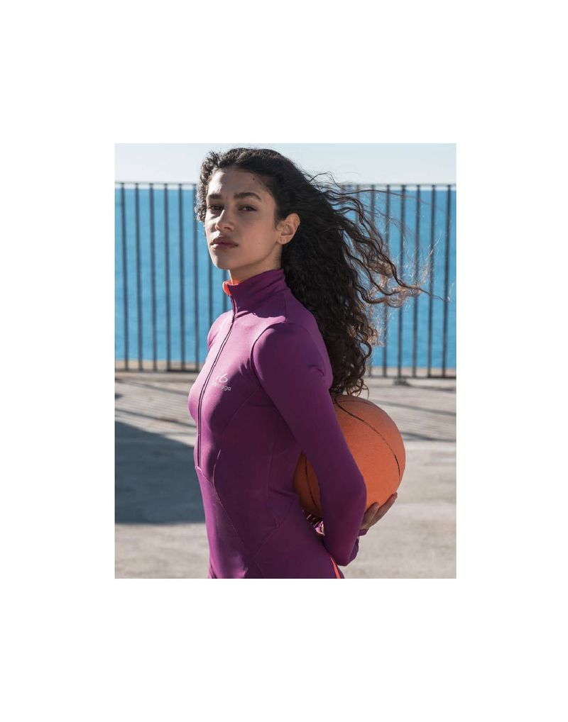 Model Tamar David wearing a purple long-sleeve athletic top and holding an orange ball behind her back near the sea.