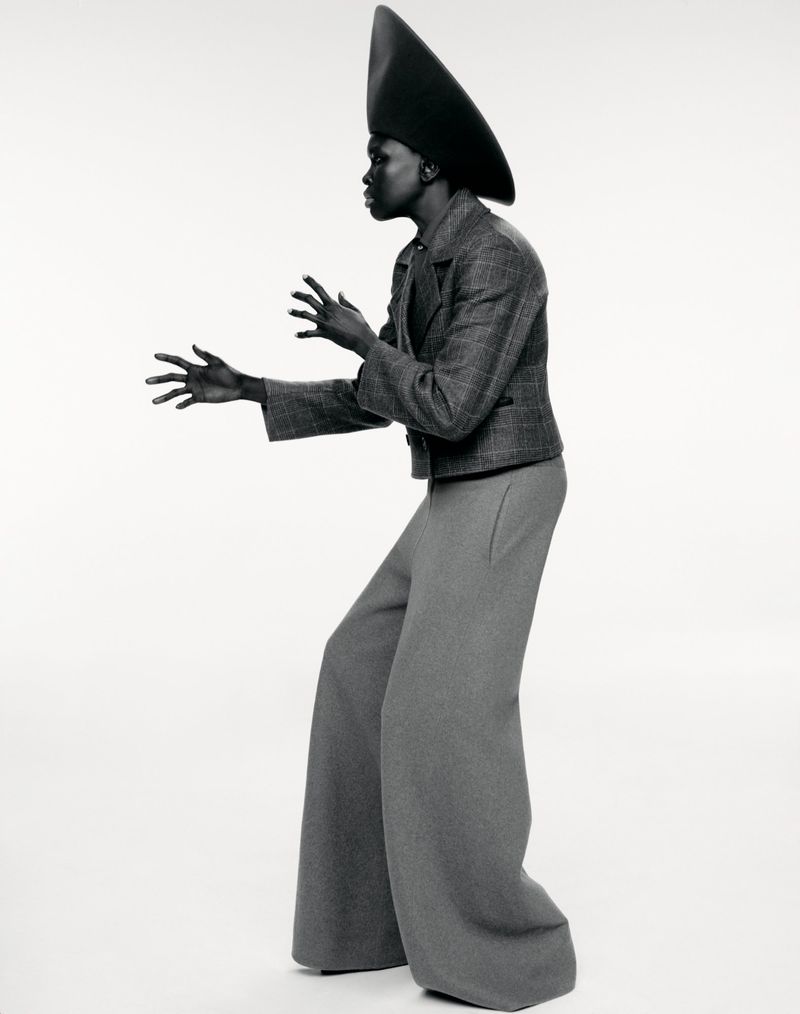 Model Alek Wek wearing a structured grey plaid jacket, wide trousers, and a tall conical hat against white