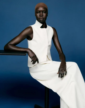 Model Alek Wek seated in a sleeveless white ensemble resting her arm on a black table against a solid blue backdrop.