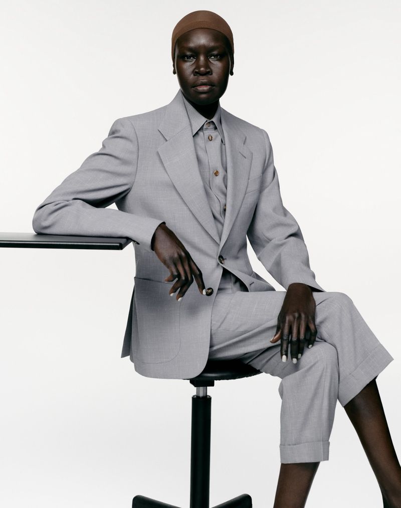 Model Alek Wek seated in a light grey tailored trouser suit and brown cap against a white background.