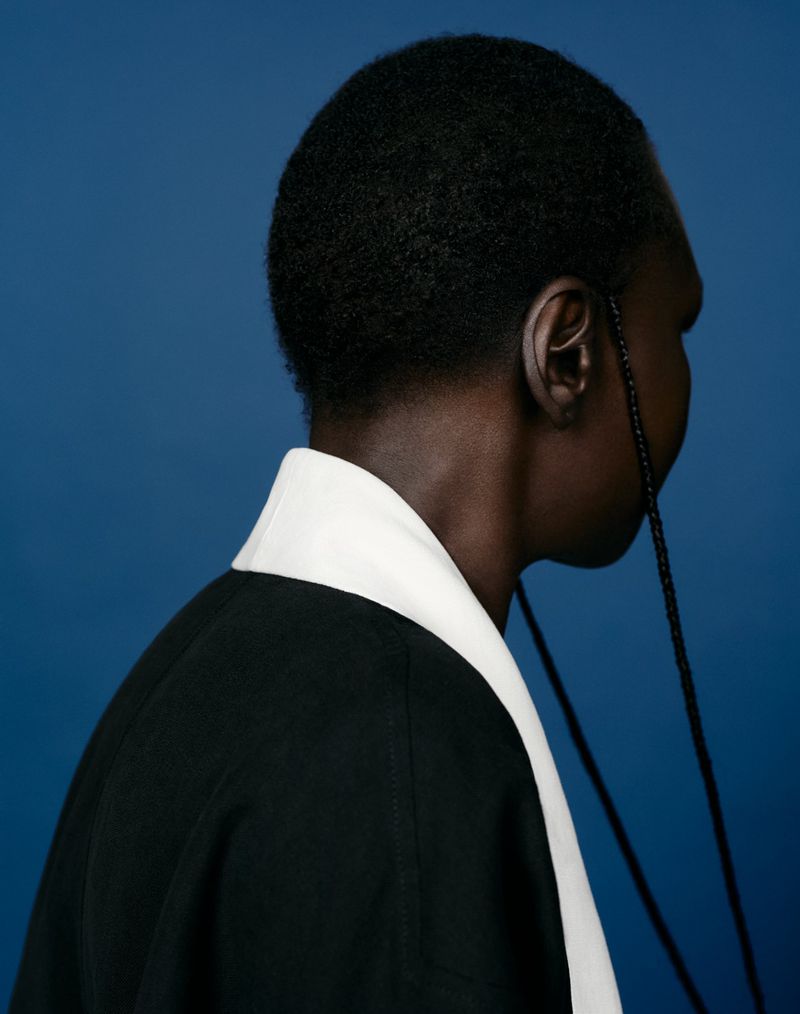 Model Alek Wek in profile wearing a black garment with white collar against a blue seamless background for CAP 74024.