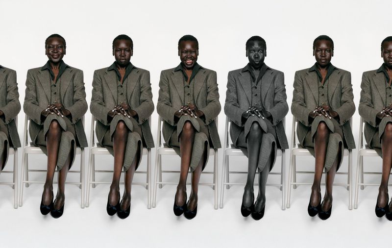 Model Alek Wek seated in repeated poses wearing a grey tailored suit and black flats across white chairs against a white background.