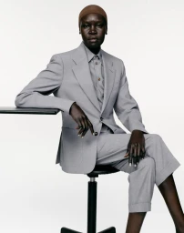 Model Alek Wek seated in a light grey tailored trouser suit and brown cap against a white background.