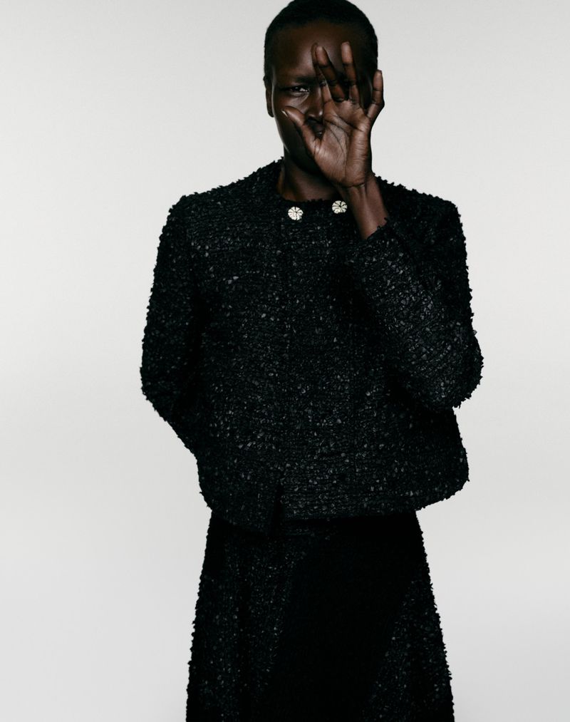 Model Alek Wek posing with a raised hand over her face in a black textured tweed jacket against a studio backdrop.