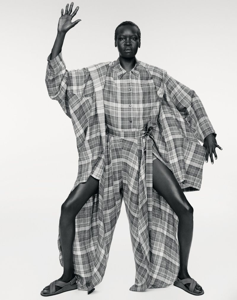 Model Alek Wek poses in an oversized plaid draped ensemble and sandals against a white studio background in black and white.