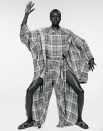 Model Alek Wek poses in an oversized plaid draped ensemble and sandals against a white studio background in black and white.