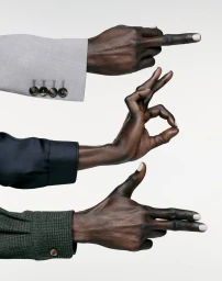 Three hands of model Alek Wek emerging from tailored jacket sleeves making gestures against a white seamless background.