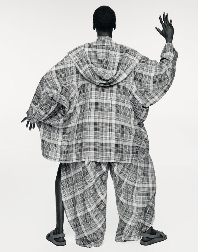 Model Alek Wek seen from behind wearing an oversized plaid hooded cape and wide-leg matching trousers against a white seamless backdrop.