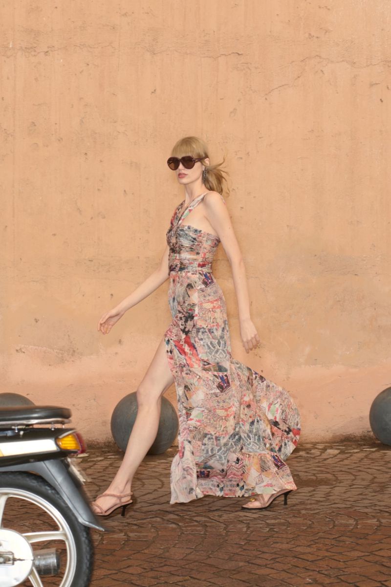 Woman in a floral dress walks near a moped. She wears sunglasses and sandals, and is posing outdoors near a peach colored wall.