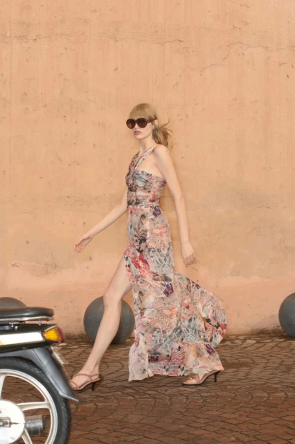 Woman in a floral dress walks near a moped. She wears sunglasses and sandals, and is posing outdoors near a peach colored wall.