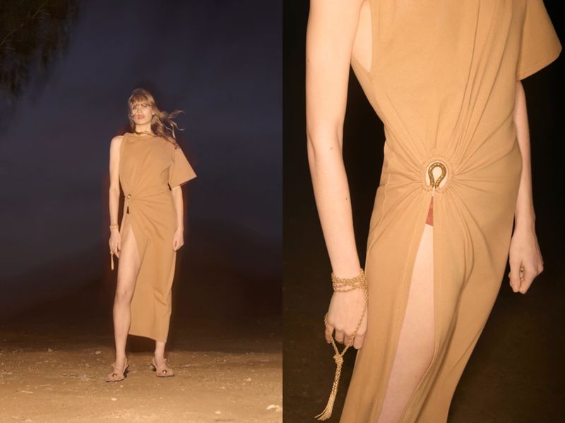 Fashion shot of a woman wearing a tan, asymmetrical dress with a thigh-high slit, standing in a dark outdoor location, accessorized with jewelry.