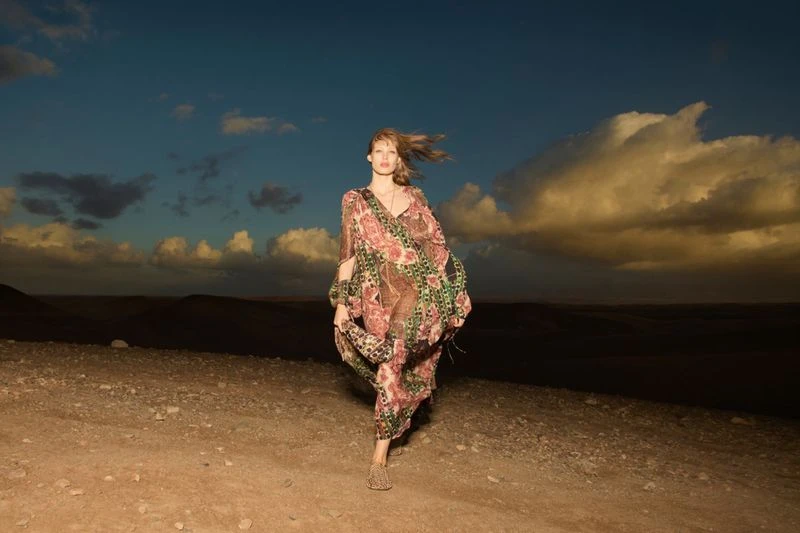 Adult woman in a patterned dress walks along a desert road under a blue sky with clouds at dusk; fashion, style, and desert landscape.