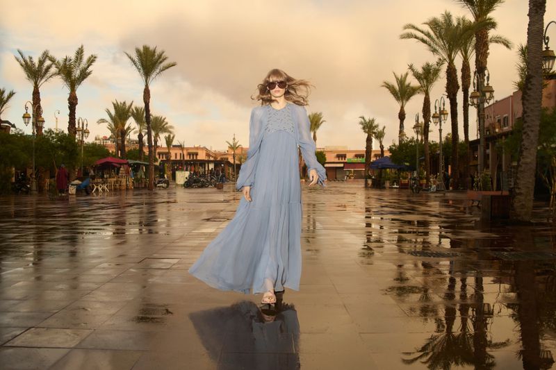 Woman in flowing blue dress and sunglasses walks on wet pavers lined with palm trees, bathed in soft, overcast light.