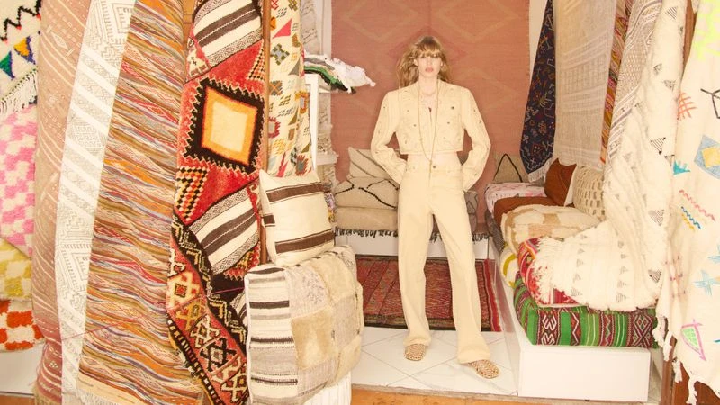 A woman in a cream-colored suit stands in front of a display of stacked rugs and textiles in a shop with neutral lighting and colors.
