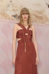 Model wearing a rust-colored ruched dress with cutout details and a long gold necklace against a textured pink and weathered wall