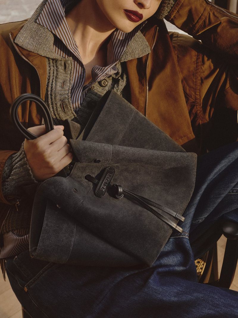 Model Agnes Wahlström seated holding a charcoal suede bucket bag wearing a brown jacket and denim jeans for Ferrari Pre-Fall 2026.