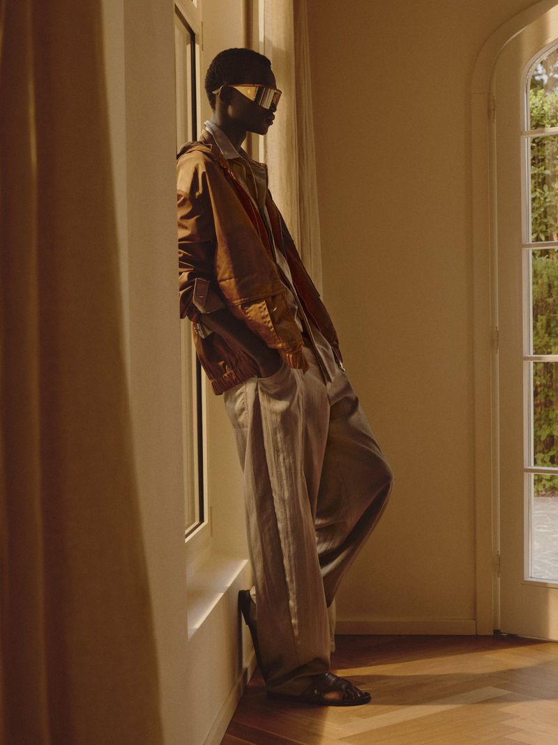 Model wearing a brown jacket, visor sunglasses, and wide-leg trousers leaning against an interior window frame.