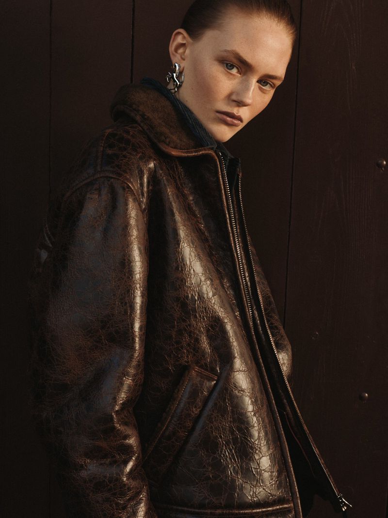 Model wearing a dark brown distressed leather jacket and silver earrings against a dark panelled wall.