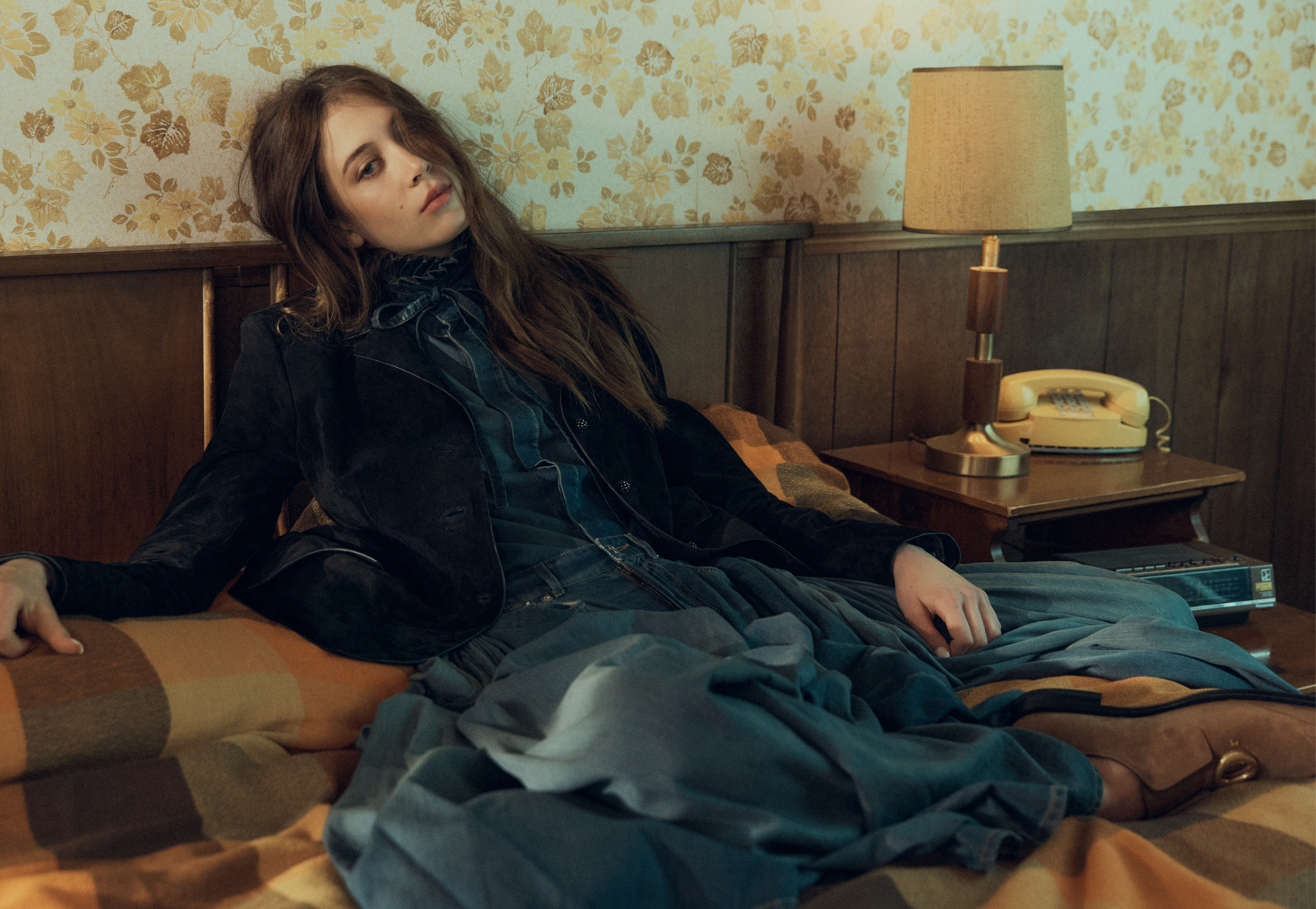 A young female model reclining on a retro bed against floral wallpaper with a bedside lamp and rotary telephone.