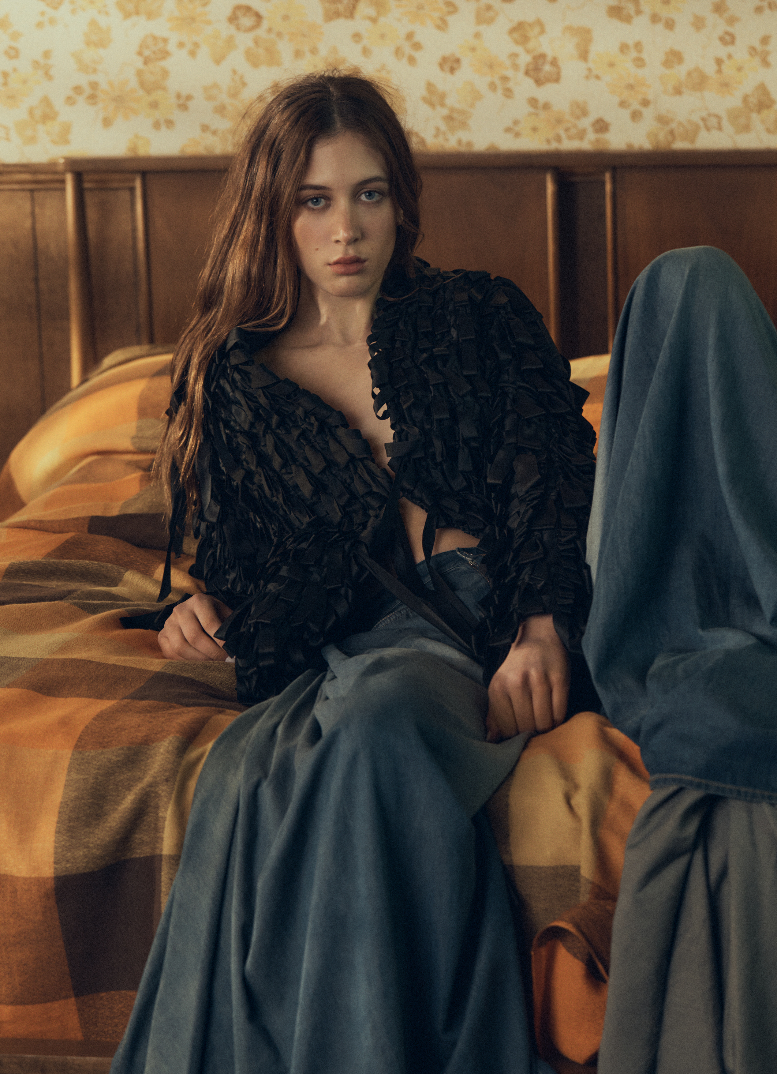 Sunday Rose Kidman Urban wearing a black textured jacket and denim skirt, seated on a bed with a plaid blanket.