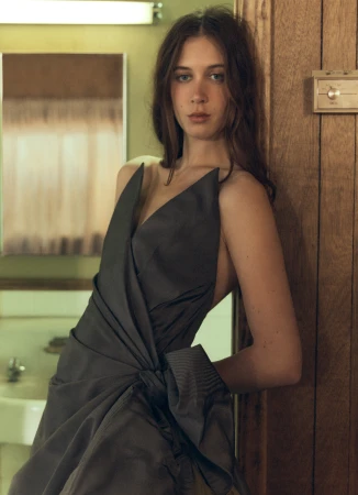 Sunday Rose wearing a structured dark charcoal draped dress while leaning against a wooden doorframe.