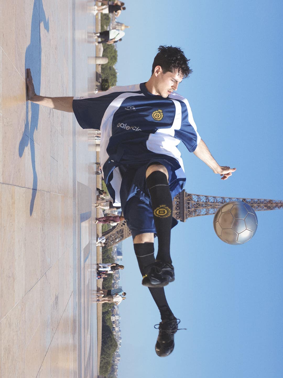 Balenciaga soccer ad campaign image featuring a male player in blue and white sportswear juggling a silver football outdoors.