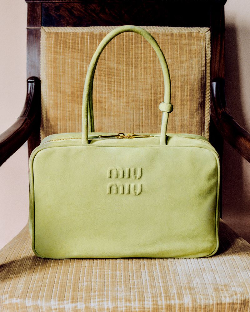 Lime green Miu Miu handbag with top handles and gold zipper, centered on a tan textured chair with a dark wood frame.