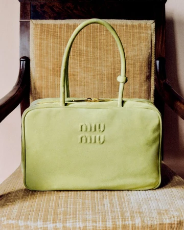 Lime green Miu Miu handbag with top handles and gold zipper, centered on a tan textured chair with a dark wood frame.
