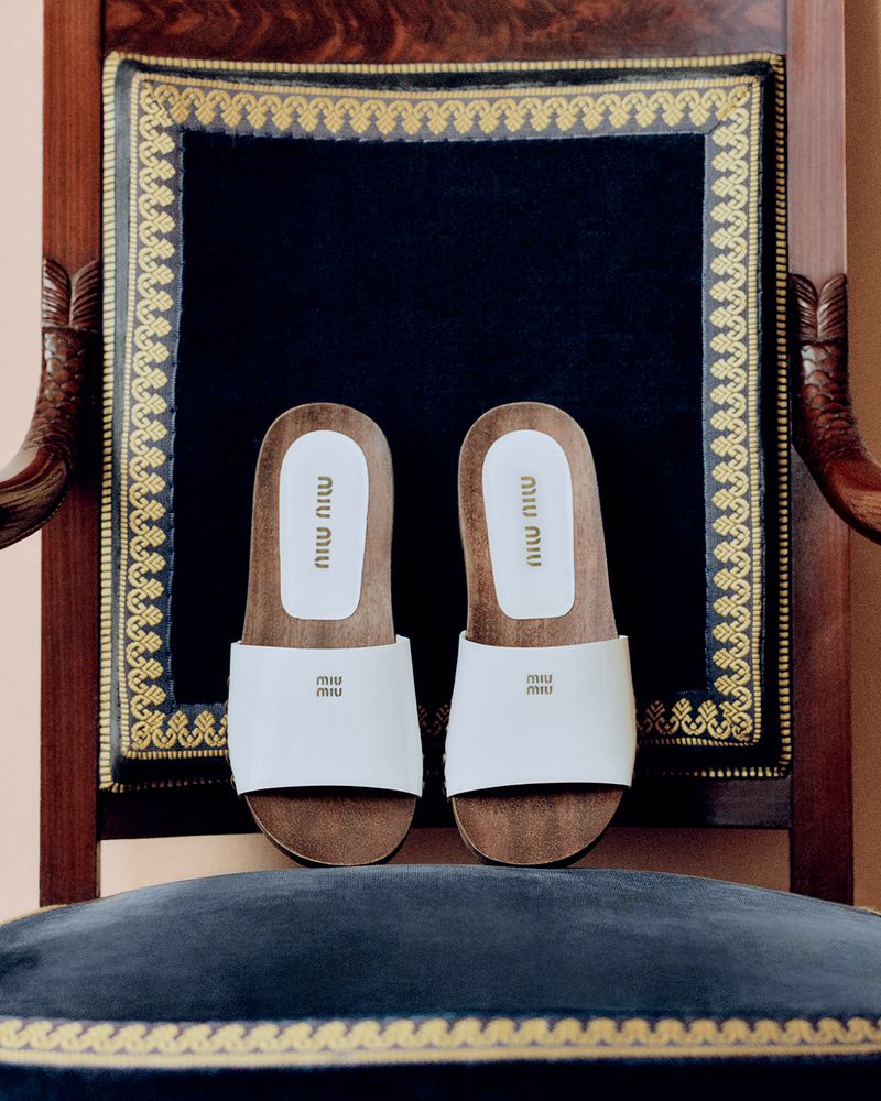 Miu Miu slide sandals showcased on an ornate, dark wood chair with plush blue velvet seating and a decorative gold trim.