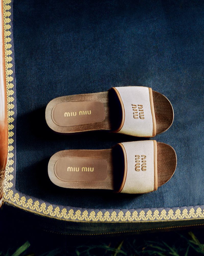 Two Miu Miu branded ivory-colored slide sandals displayed on a dark blue textured surface, framed by a decorative gold trim. 