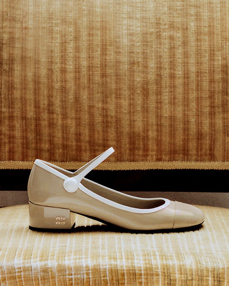 Beige patent leather Miu Miu Mary Jane shoe with white trim and strap, displayed on a striped fabric surface against a tan curtain.