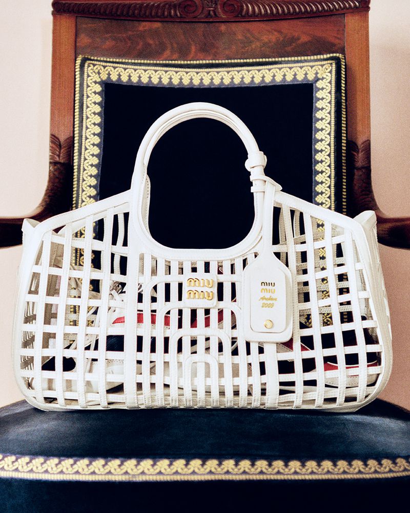 A white Miu Miu basket-weave handbag is displayed on a blue velvet cushioned chair against a dark ornate backdrop. Fashion product photography.