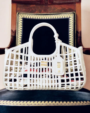 A white Miu Miu basket-weave handbag is displayed on a blue velvet cushioned chair against a dark ornate backdrop. Fashion product photography.