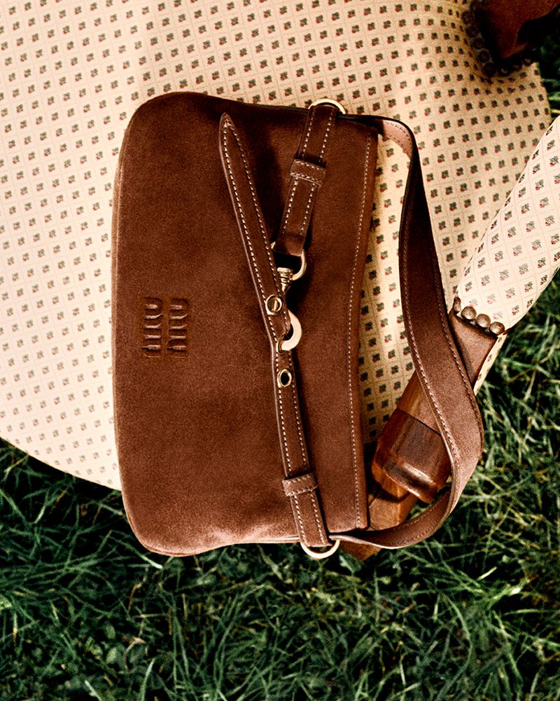 Brown suede purse with strap and silver hardware sitting on a patterned chair cushion with a grass background in an outdoor setting.