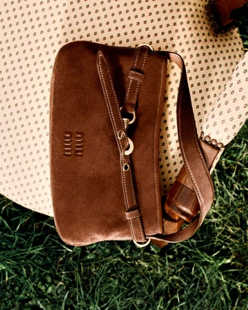 Brown suede purse with strap and silver hardware sitting on a patterned chair cushion with a grass background in an outdoor setting.