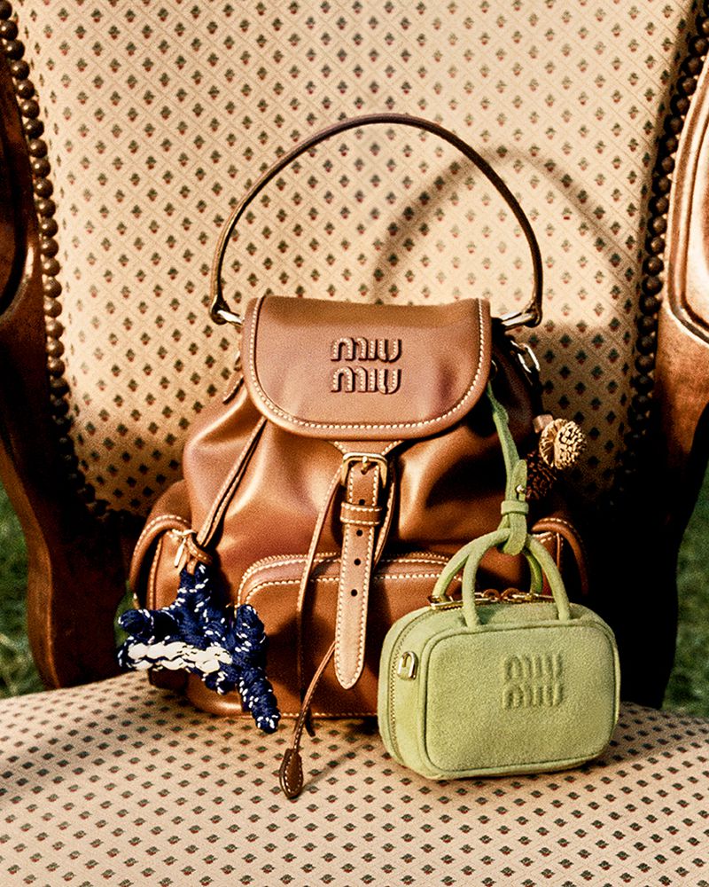 Two Miu Miu brand handbags, one brown leather backpack and a light green, small square bag, are styled on an ornate chair.