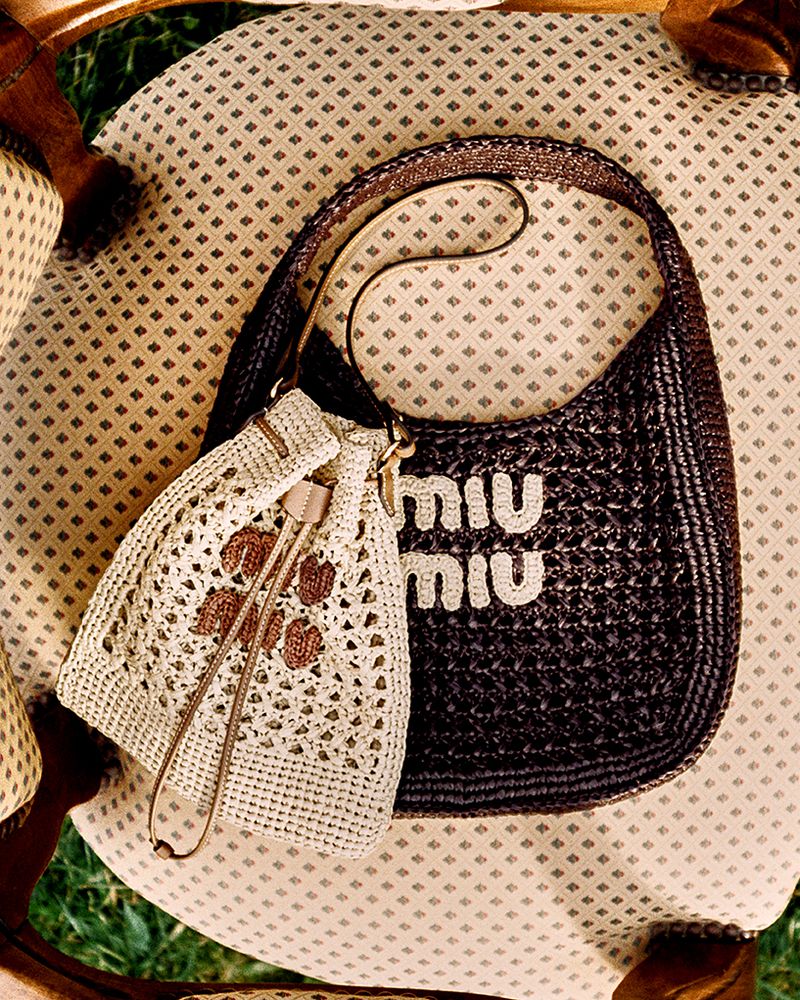 Still life shot of a dark brown woven handbag with the MIU MIU logo, and a light beige crocheted bucket bag, placed on a patterned chair.